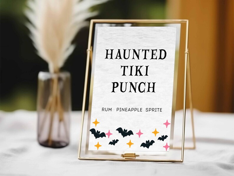 May include: A gold framed sign with the words "HAUNTED TIKI PUNCH" in black. The sign also says "RUM PINEAPPLE SPRITE" below the title. The bottom of the sign has black bats, pink stars, and orange stars.