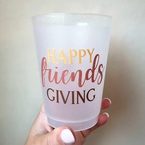 Peut inclure: Un gobelet en plastique givr&eacute; avec les mots "HAPPY friends GIVING" en lettres dor&eacute;es et or rose. La tasse est tenue par une main avec des ongles peints.
