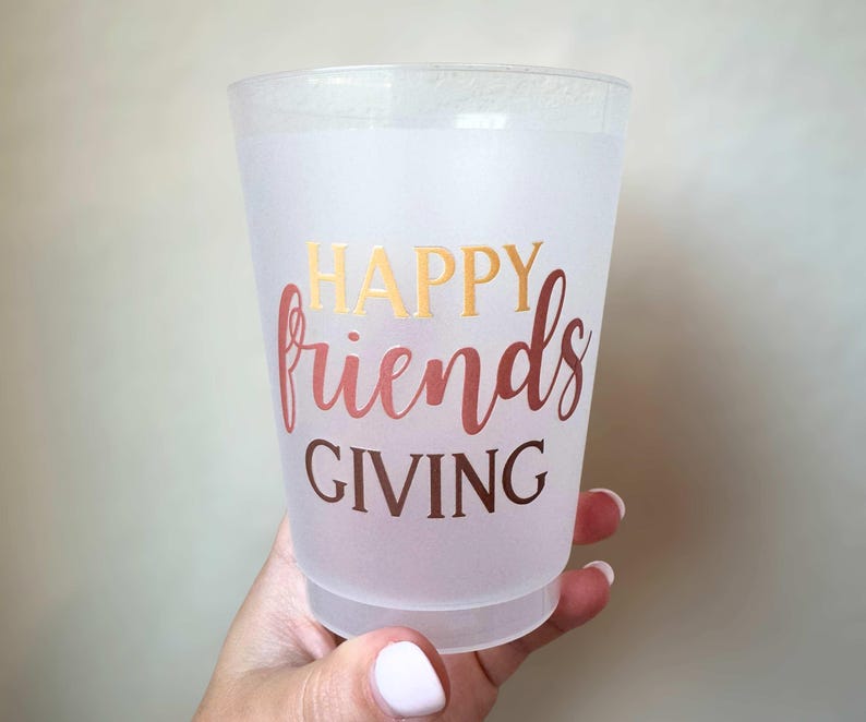Peut inclure: Un gobelet en plastique givr&eacute; avec les mots "HAPPY friends GIVING" en lettres dor&eacute;es et or rose. La tasse est tenue par une main avec des ongles peints.