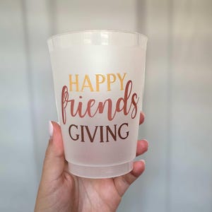 Peut inclure: Un gobelet en plastique givr&eacute; avec les mots "HAPPY friends GIVING" en or et en script rouge. La tasse est tenue par une main, et l'arri&egrave;re-plan est gris clair.