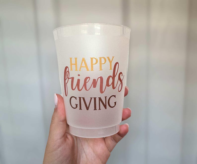 Peut inclure: Un gobelet en plastique givr&eacute; avec les mots "HAPPY friends GIVING" en or et en script rouge. La tasse est tenue par une main, et l'arri&egrave;re-plan est gris clair.