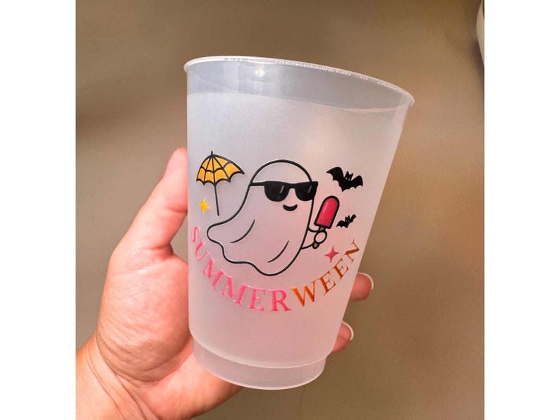 May include: A frosted plastic cup with a "SUMMERWEEN" design. The design features a cartoon ghost wearing sunglasses and holding a popsicle, with a yellow umbrella and bats. The text is in pink and gold.