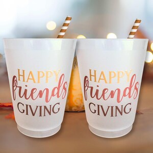 Peut inclure: Deux gobelets en plastique blancs avec l'inscription "HAPPY friends GIVING" en lettres dor&eacute;es et rouges. Chaque gobelet est orn&eacute; d'une paille ray&eacute;e marron et blanche. L'arri&egrave;re-plan est flou avec des couleurs d'automne.