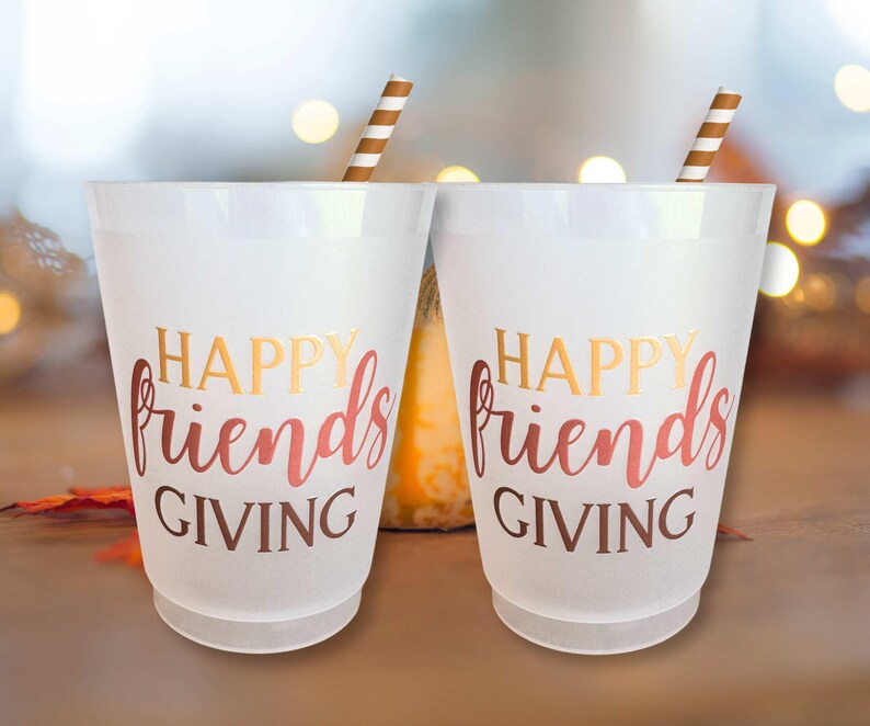 Peut inclure: Deux gobelets en plastique blancs avec l'inscription "HAPPY friends GIVING" en lettres dor&eacute;es et rouges. Chaque gobelet est orn&eacute; d'une paille ray&eacute;e marron et blanche. L'arri&egrave;re-plan est flou avec des couleurs d'automne.