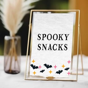 May include: A gold framed sign with the words "SPOOKY SNACKS" in black. The bottom of the sign is decorated with black bats, orange stars, and pink stars. The sign is on a white surface.