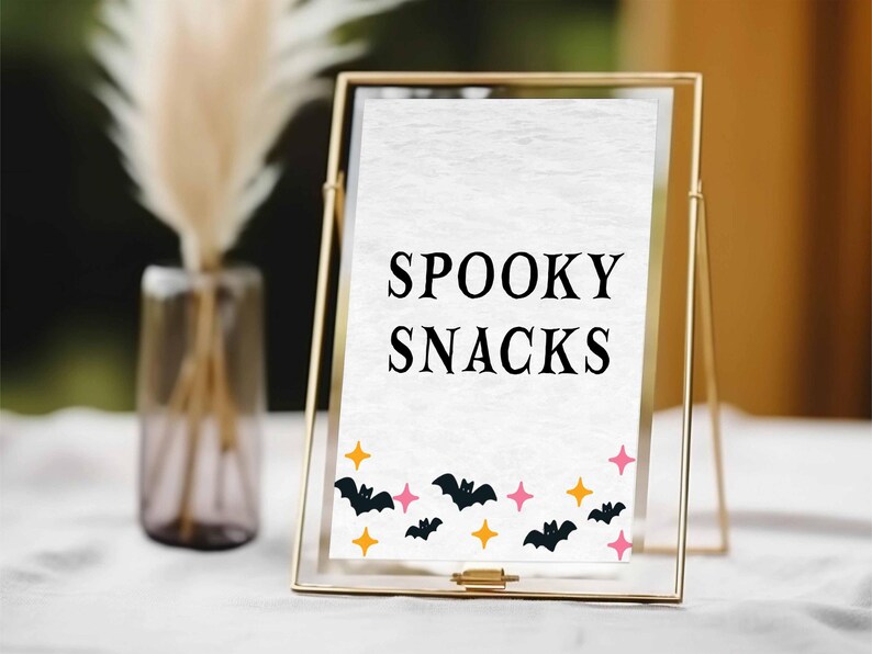 May include: A gold framed sign with the words "SPOOKY SNACKS" in black. The bottom of the sign is decorated with black bats, orange stars, and pink stars. The sign is on a white surface.