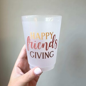 Peut inclure: Un gobelet en plastique transparent avec les mots "HAPPY friends GIVING" en lettres dor&eacute;es et or rose. Le gobelet est tenu par une main avec du vernis &agrave; ongles rose clair.