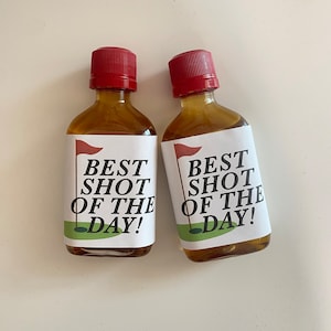 May include: Two small glass bottles with red caps and labels that say "Best Shot of the Day!" with a golf flag and green golf course graphic.