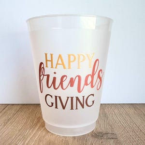 Peut inclure: Un gobelet en plastique transparent avec les mots "HAPPY friends GIVING" en lettres dor&eacute;es et rouge-orang&eacute;. Le gobelet a un aspect givr&eacute; et repose sur une surface en bois.