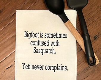 May include: White kitchen towel with black text that reads "Bigfoot is sometimes confused with Sasquatch. Yeti never complains."