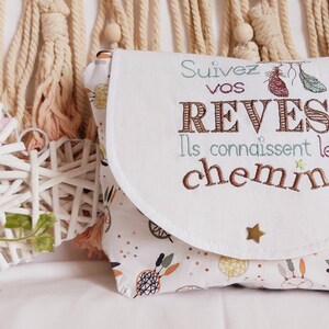 May include: White fabric pouch with a flap closure. The pouch has a white background with a colorful pattern of dreamcatchers and feathers. The flap has a white background with the text "Suivez vos reves Ils connaissent le chemin" embroidered in brown, blue, and pink thread.