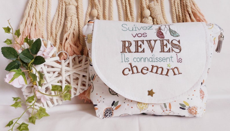 May include: White fabric pouch with a flap closure. The pouch has a white background with a colorful pattern of dreamcatchers and feathers. The flap has a white background with the text "Suivez vos reves Ils connaissent le chemin" embroidered in brown, blue, and pink thread.