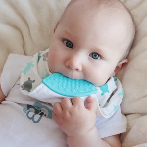 Puede incluir: Un bebé con un babero blanco con un patrón azul y gris está masticando un mordedor de silicona azul. El mordedor tiene una superficie texturizada.