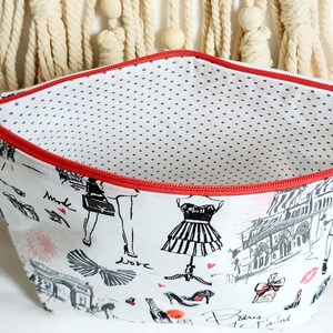 May include: A white zippered pouch with a red zipper and a black and white print of Parisian themed illustrations. The inside of the pouch is lined with white fabric with black polka dots.