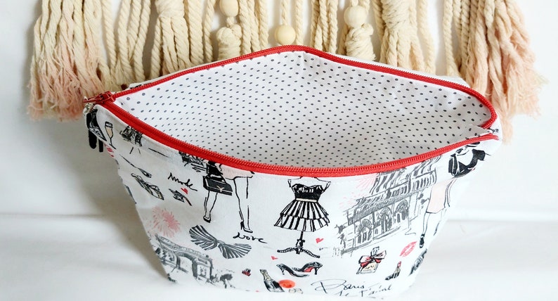 May include: A white zippered pouch with a red zipper and a black and white print of Parisian themed illustrations. The inside of the pouch is lined with white fabric with black polka dots.
