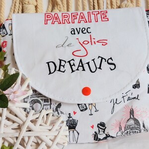 May include: White fabric makeup bag with a red button closure. The bag has a flap with black embroidery that reads "PARFAITE avec de jolis DEFAUTS". The bag is printed with black and white illustrations of Parisian landmarks and fashion.