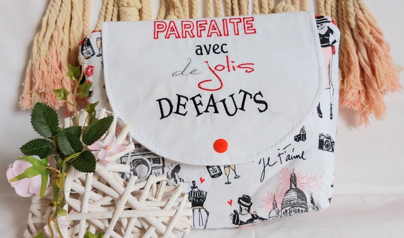 May include: White fabric makeup bag with a red button closure. The bag has a flap with black embroidery that reads "PARFAITE avec de jolis DEFAUTS". The bag is printed with black and white illustrations of Parisian landmarks and fashion.