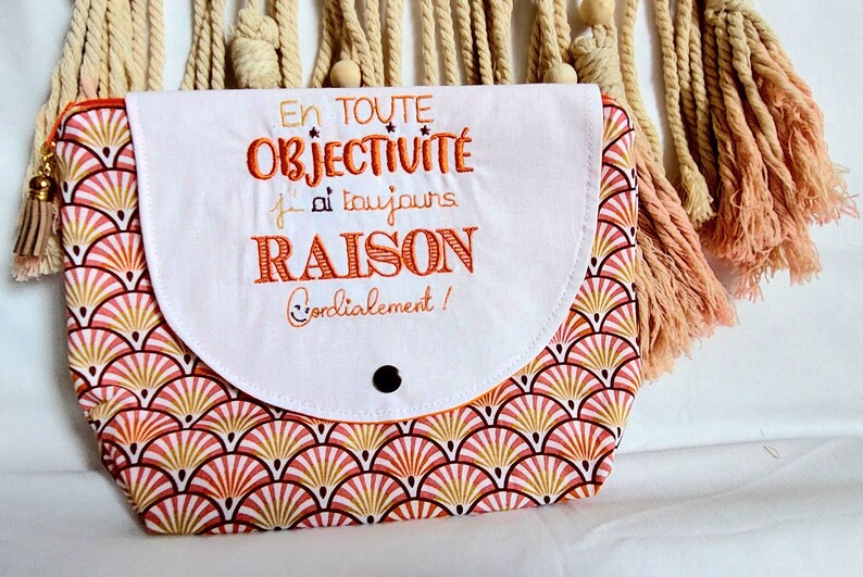 May include: White zippered pouch with a pink and gold geometric pattern. The pouch has a white flap with the text "En toute objectivit&eacute; j'ai toujours RAISON Cordialement!" embroidered in orange.