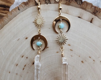 May include: Gold earrings with a crescent moon, starburst, and clear quartz crystal drop. The earrings are on a wooden surface.