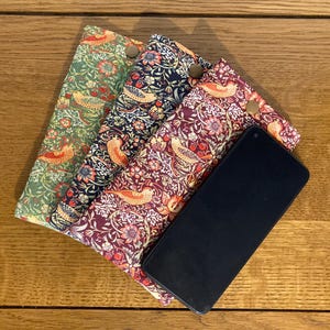 May include: Three fabric phone cases with a floral pattern and birds. The cases are in shades of green, blue, and red. A black smartphone is lying on top of the cases.