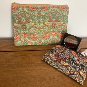 May include: Two patterned cosmetic bags, one rectangular with a zip, the other open, displaying a compact mirror and lipstick. The bags feature a floral and bird design in green, red, and yellow.
