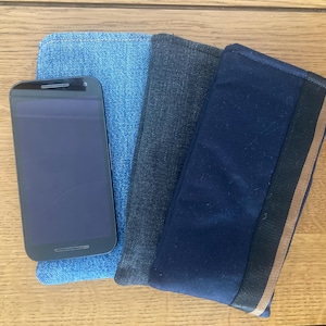 May include: A smartphone sits on a stack of three denim fabric cases. The cases are in varying shades of blue, with one featuring a black and gold stripe. The items are arranged on a wooden surface.