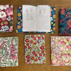 May include: Several fabric earring holders with floral designs in shades of red, blue, and purple. Some are open, revealing stud earrings. The earrings are silver and gold coloured. The holders are arranged on a wooden surface.