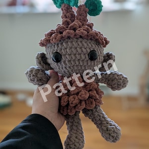May include: A crocheted amigurumi toy with a brown body, brown hair, and green leaves on top. The toy has large black eyes and a small mouth.