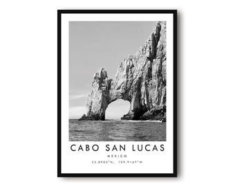 May include: Black and white framed print featuring a scenic view of Cabo San Lucas, Mexico. The image showcases a natural rock archway over the ocean. Text at the bottom reads "CABO SAN LUCAS, MEXICO" with coordinates.