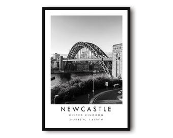 May include: Black and white framed print featuring a photograph of the Tyne Bridge in Newcastle, United Kingdom. The image includes the bridge, river, and surrounding buildings. The word "NEWCASTLE" is printed at the bottom, along with the country and coordinates.