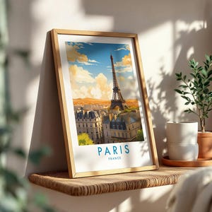 Op de afbeelding: Een ingelijste kunstdruk met een gestileerde illustratie van de Eiffeltoren in Parijs, Frankrijk. De afbeelding toont een stadsgezicht onder een blauwe lucht met gouden wolken, met de tekst PARIS FRANCE in het blauw onderaan de witte rand.
