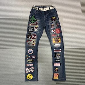 Peut inclure: Jean en denim bleu avec de nombreux écussons colorés et une ceinture blanche. Les écussons comprennent du texte et des graphiques de dessins animés. Le jean a une coupe droite et un aspect usé. La ceinture a des clous argentés.