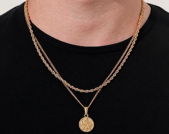 May include: A layered gold-tone necklace set featuring a shorter rope chain and a longer, thinner chain with a circular pendant. The pendant displays a detailed embossed figure on its face. The jewelry is worn against a plain black t-shirt.