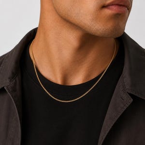May include: A thin gold chain necklace with a classic curb link design resting against a black t-shirt and a dark gray collared shirt. The metallic yellow gold links are uniform in size and shape, creating a simple and refined jewelry piece.