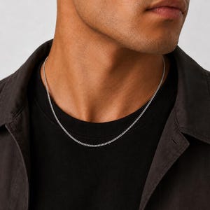 May include: A thin, silver-toned metal chain necklace with small, interlocking links worn around the neck. The necklace rests against a plain black t-shirt, which is layered underneath a dark charcoal gray button-down shirt with a visible collar.