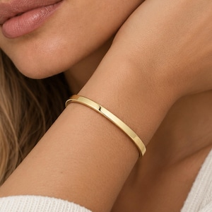 May include: A polished gold-tone metal bangle bracelet with a smooth, rounded surface and a continuous circular design. The bracelet features a reflective, high-shine finish that catches the light, creating a clean and simple aesthetic on the wrist.