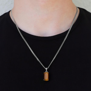 May include: A silver chain necklace with a rectangular, amber-coloured pendant. The pendant is a warm, golden-brown hue. The necklace is worn against a black top.