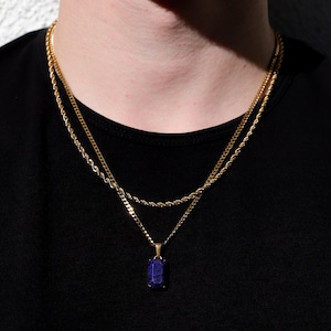 May include: Gold-toned chain necklaces layered on a black shirt. The top chain is a curb link, and the bottom chain is a rope style. A gold pendant with a rectangular, dark blue gemstone hangs from the bottom chain.