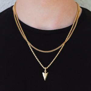 Puede incluir: Un collar de cadena de oro con un colgante de flecha de oro. El collar está superpuesto con una cadena de oro más gruesa.
