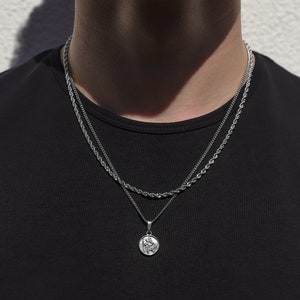 Puede incluir: Un collar de cadena de plata con un colgante redondo. El collar está superpuesto a una segunda cadena de plata más fina.