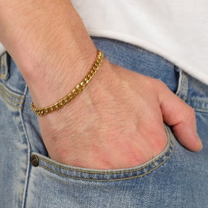 Peut inclure: Un bracelet en cha&icirc;ne dor&eacute;e au poignet d'une personne. Le bracelet est fait d'une cha&icirc;ne &eacute;paisse de style curb.