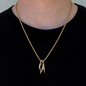 May include: A gold-colored rope chain necklace with two pendants. One pendant is a curved horn shape, and the other is a hand gesture. The necklace is worn against a black shirt.
