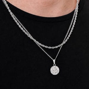 Puede incluir: Un conjunto de collares plateados en capas que incluye una cadena de cuerda más corta y una cadena más larga con un colgante circular. El colgante redondo muestra una figura grabada detallada y ambas cadenas son de metal pulido.