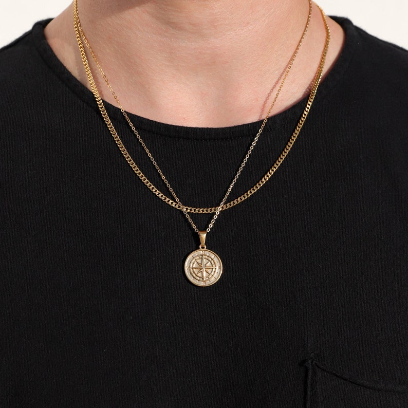 May include: A gold chain necklace with a compass pendant. The chain is layered with a thinner chain.