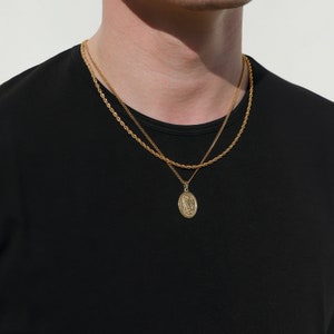 May include: Two gold chains with a pendant featuring a religious figure. The chains are layered and the pendant is oval shaped.