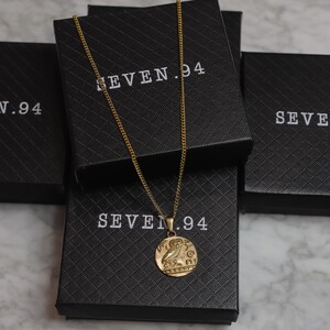 May include: Gold chain necklace with a round pendant featuring an owl design. The necklace is displayed in a black box with the text "SEVEN.94" printed on it.