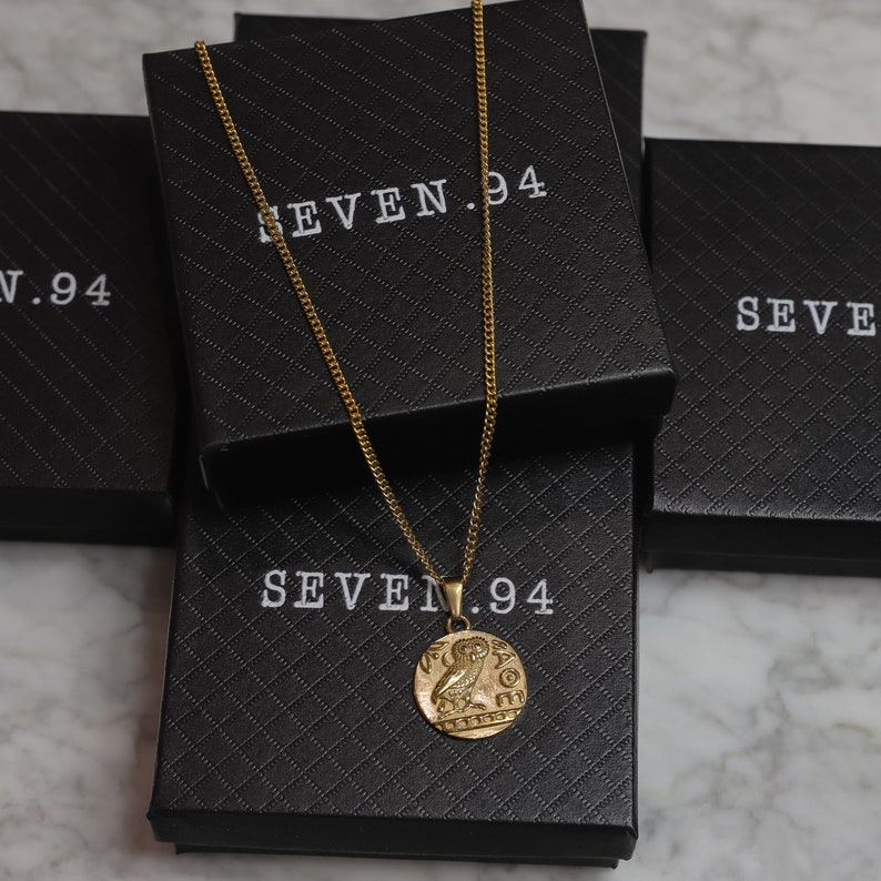 May include: Gold chain necklace with a round pendant featuring an owl design. The necklace is displayed in a black box with the text "SEVEN.94" printed on it.