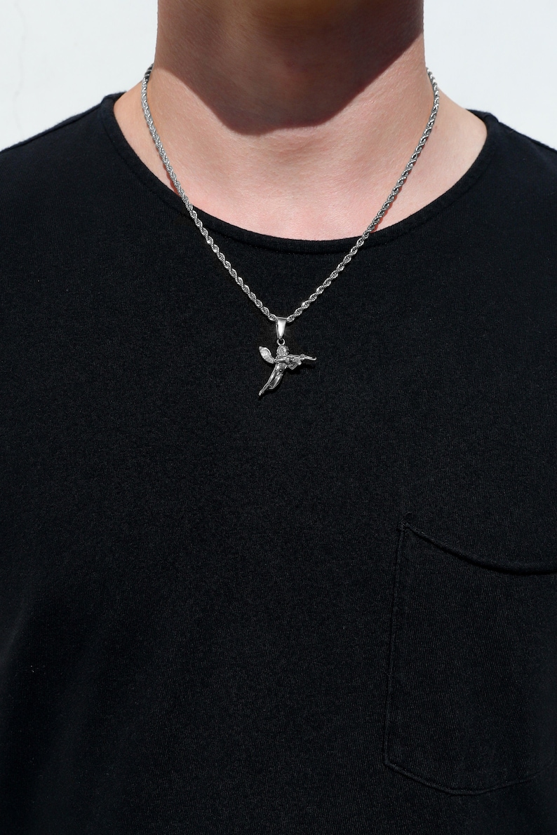 Puede incluir: Un collar de cadena de plata con un colgante de &aacute;ngel de plata. El collar lo lleva una persona que lleva una camiseta negra.