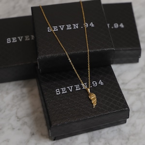 Puede incluir: Un collar de cadena dorada con un peque&ntilde;o colgante de ala dorada. El collar se muestra en una caja negra con el texto "SEVEN.94" impreso en &eacute;l.
