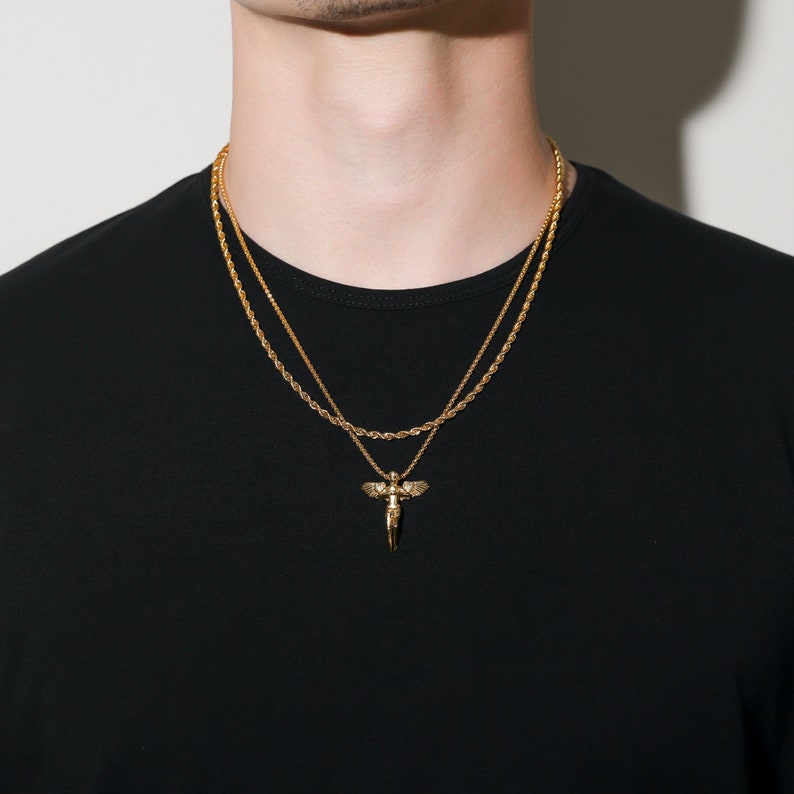 May include: A gold chain necklace with a gold angel pendant. The necklace is layered with a second, thinner gold chain.